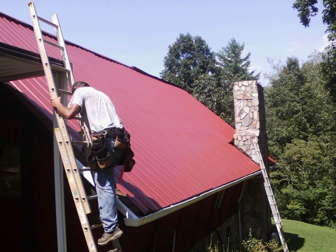 Roofer on a ladder beside a red metal roof and stone chimney — Soffit & Fascia Repair by Roofing Brothers in Sherrill